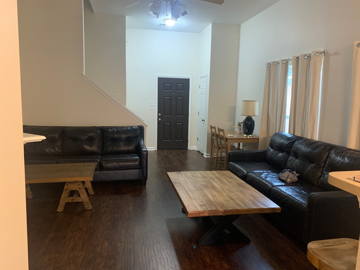 A spacious living area is seen with two black leather couches positioned around a wooden coffee table. Natural light enters through large windows, while light curtains enhance the bright ambiance. A dining table with chairs is visible, along with a staircase in the background.