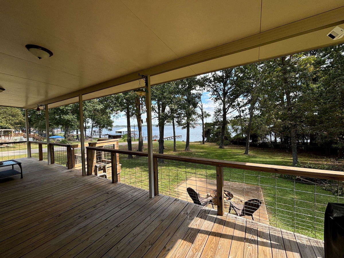 A covered deck with a wooden floor overlooks a serene lawn and Lake Palestine. Several seating areas are visible, including chairs arranged around a fire pit. Trees provide shade, while the distant shoreline and docks can be seen, enhancing outdoor relaxation.