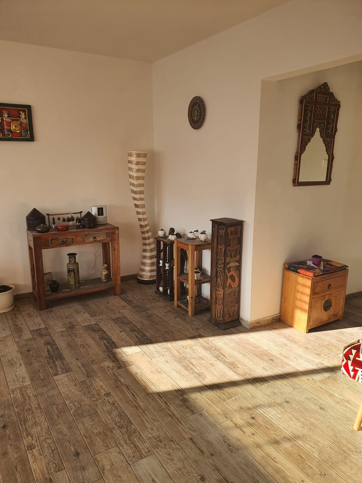 A bright room is highlighted by natural light streaming through a window, illuminating wooden flooring. Decorative elements include a tall, slender lamp, wooden furniture with intricate designs, and a large mirror. A collection of small artifacts is displayed on a console table.