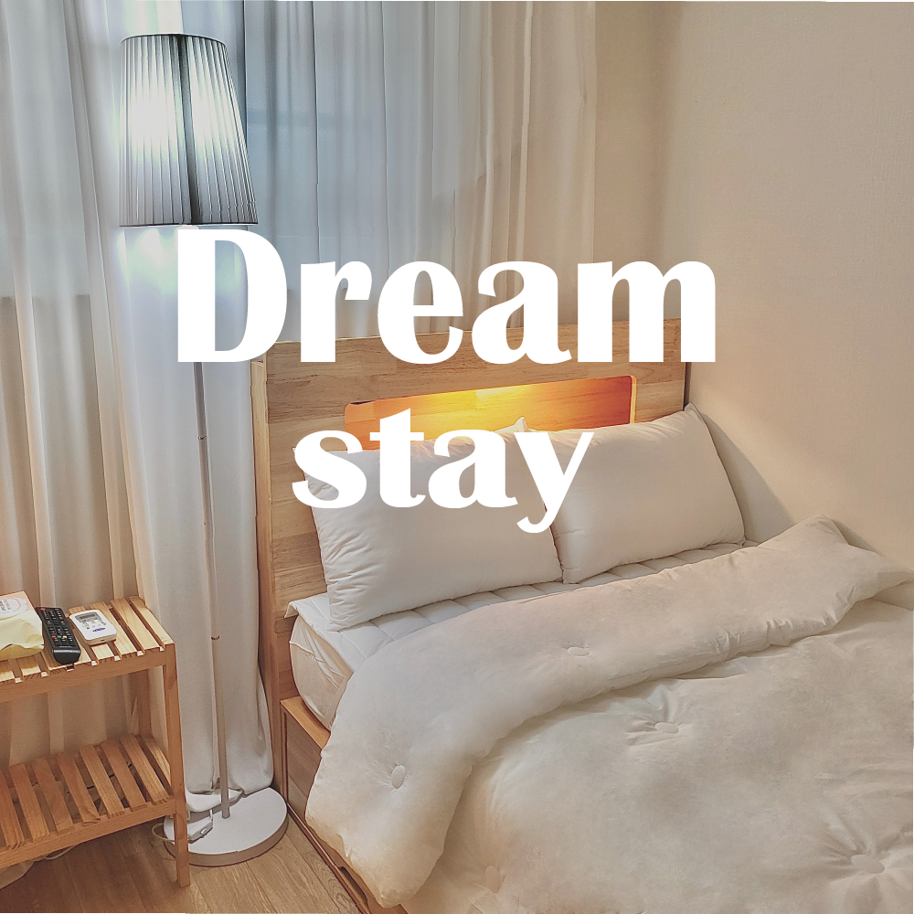 A cozy sleeping area is furnished with two spacious beds, neatly arranged with crisp white bedding. A bedside table holds a lamp, while soft light filters through sheer curtains. A wooden shelf completes the serene ambiance of the room.