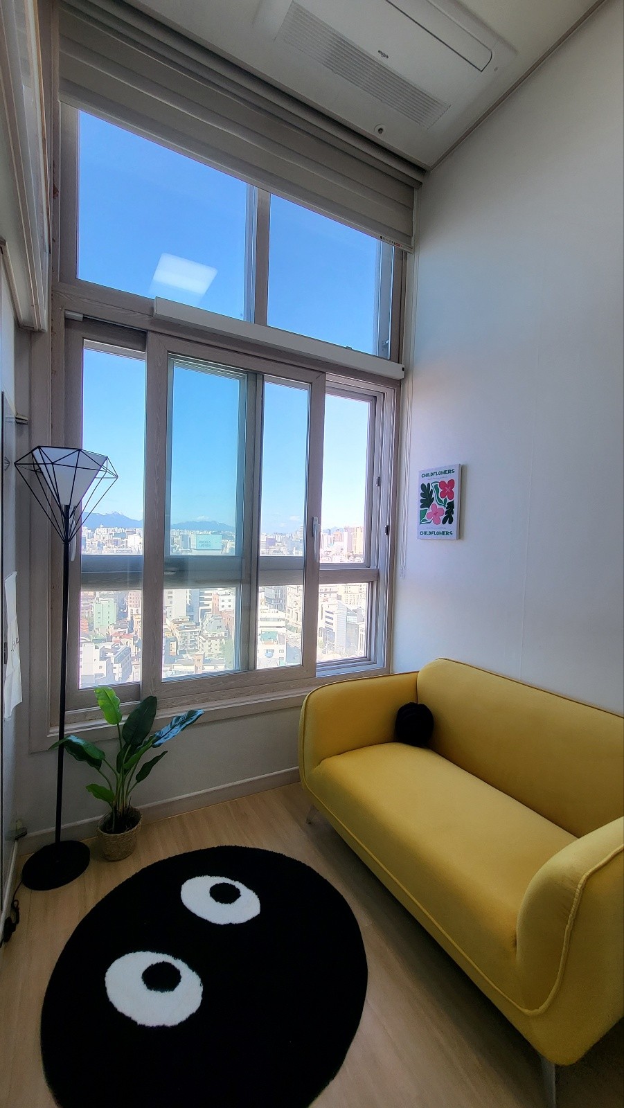A corner lounge area features a vibrant yellow sofa, positioned next to a small indoor plant. Natural light streams through a large window with multiple panes, offering a glimpse of the city skyline. A round black rug with a unique design adds character to the space.