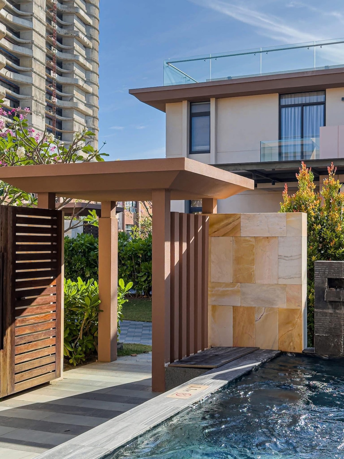 An elegant outdoor area features a private pool bordered by landscaped greenery. A contemporary shelter with a sloped roof provides a shaded entryway, while tall buildings are visible in the background, illustrating an urban vibe.