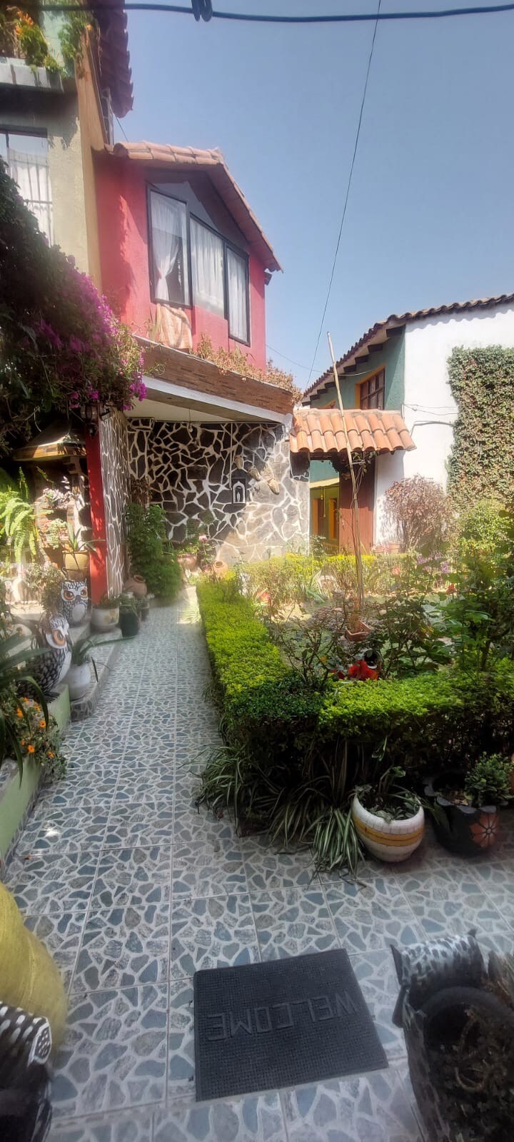 A charming pathway is lined with a variety of plants and decorative pots, leading to a vibrant building facade. Colorful flowers cascade from the upper levels, while stone walls provide texture. A 'WELCOME' mat is positioned at the entrance, inviting guests into the area.