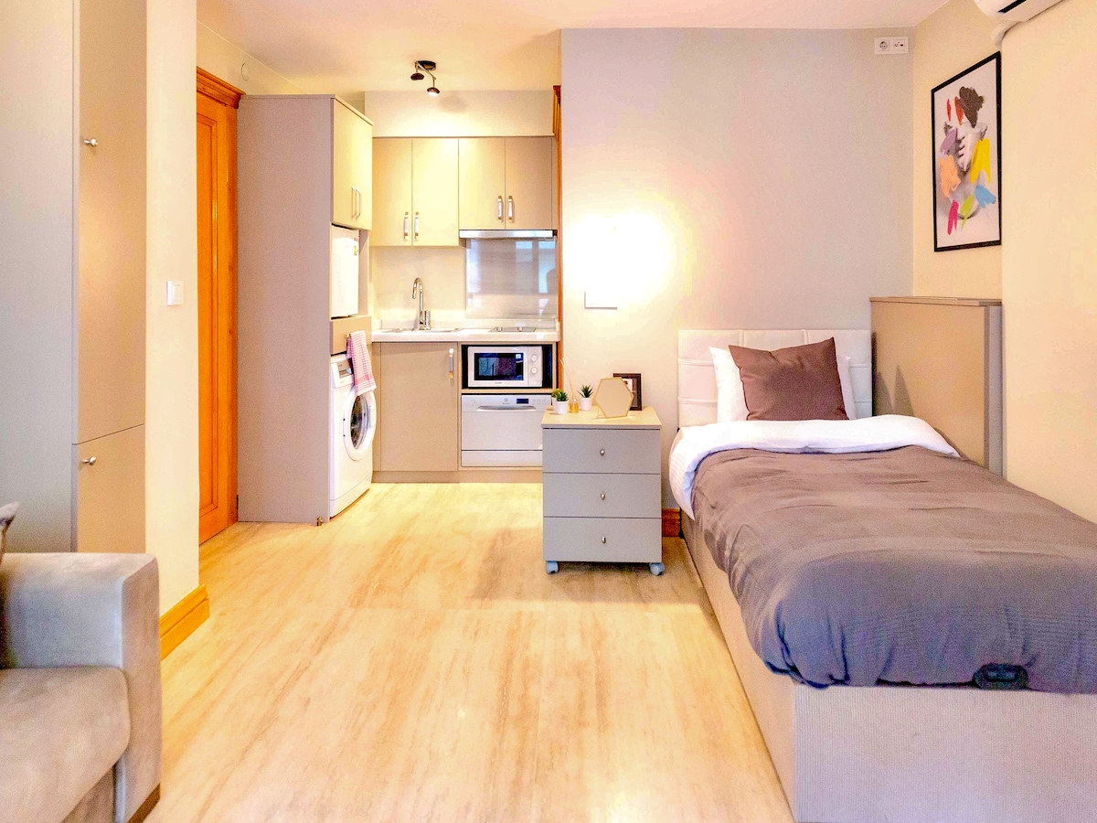 A compact studio layout is presented, featuring a spacious bed alongside a stylish sofa. The kitchenette is equipped with modern appliances, including a microwave and washing machine. Light-colored walls and a warm tone create a bright, welcoming environment, complemented by smart storage solutions.