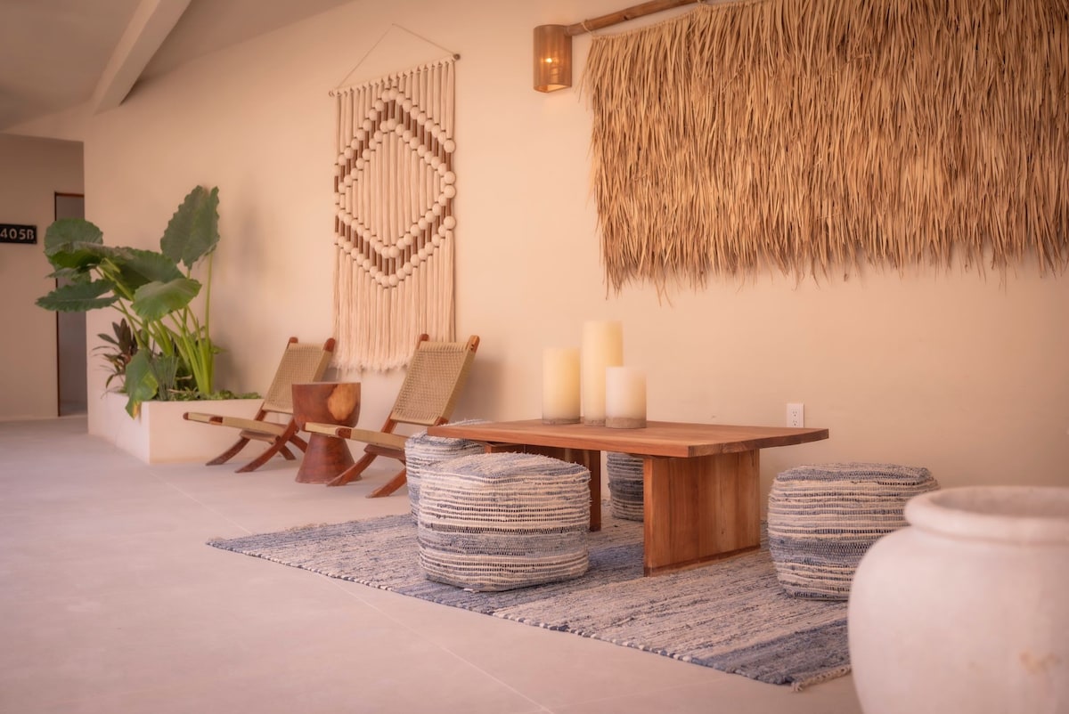 A cozy lounge area is showcased with natural decor elements, including woven wall art and a calming palette. A wooden table is complemented by low seating arrangements with round cushions, surrounded by lush green plants that add a touch of nature to the space.