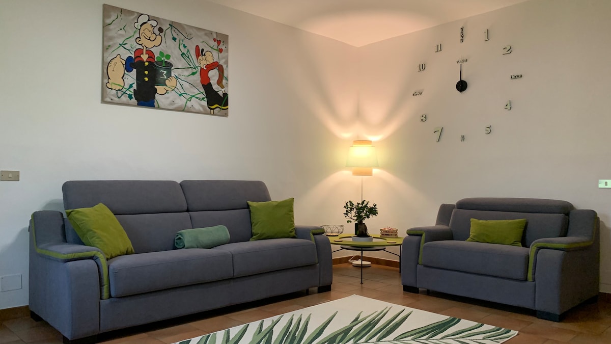 The living area features a pair of gray sofas accented with green cushions. A contemporary clock hangs on the wall above, complementing the soft lighting provided by a nearby lamp. A decorative rug with a leafy design adds a touch of warmth to the space.