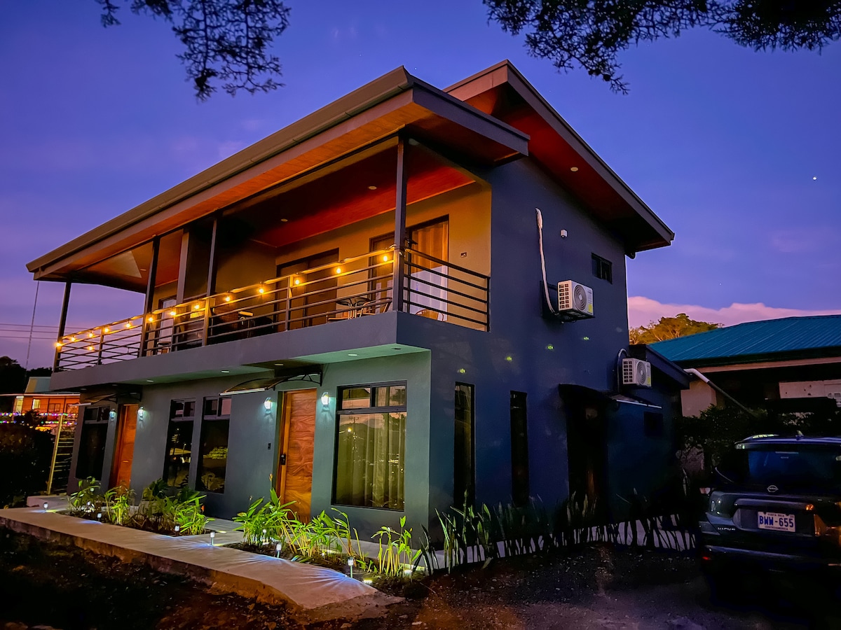 A modern two-story building is illuminated by exterior lights at dusk, highlighting its sleek design. A large balcony with railing extends across the upper floor. Surrounding greenery complements the structure, while a parking area is visible in front, accommodating vehicles.