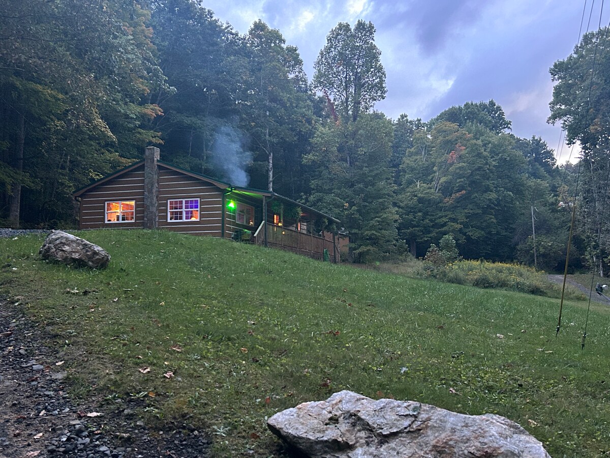 A charming cabin is set on a grassy area, surrounded by trees. Soft light glows from the windows, while a gentle plume of smoke rises from the chimney. The inviting porch is visible, extending around the structure amid the peaceful natural surroundings.