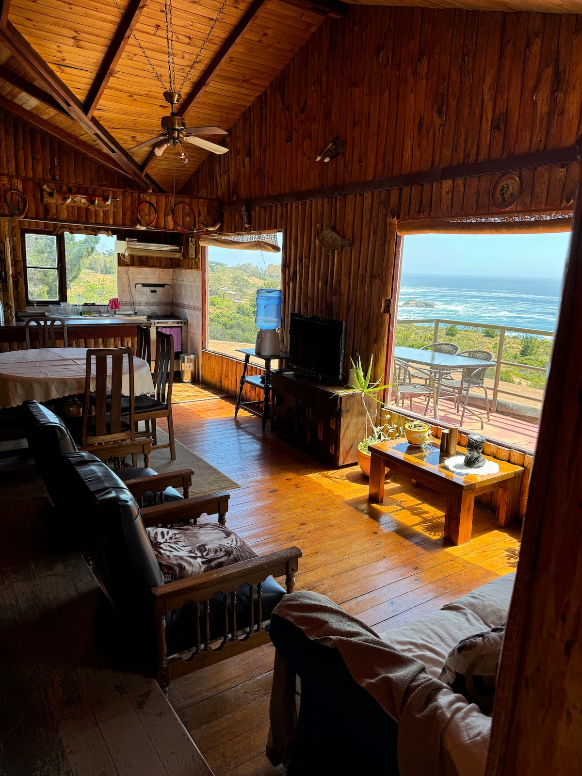 A spacious living area features wooden walls and high ceilings, complemented by large windows that provide an ocean view. Comfortable seating arrangements are placed thoughtfully, and a dining table is visible. Sunlight illuminates the warm wooden floors, enhancing the cozy atmosphere of the cabin.