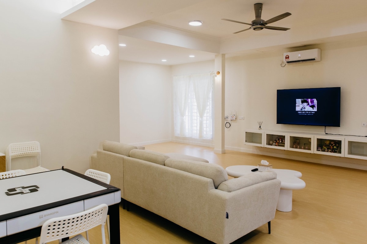 A modern living area is featured, showcasing a spacious beige sofa facing a wall-mounted television. A contemporary coffee table occupies the center. Natural light is invited through sheer curtains, enhancing the clean lines of the room, which is finished in neutral tones.