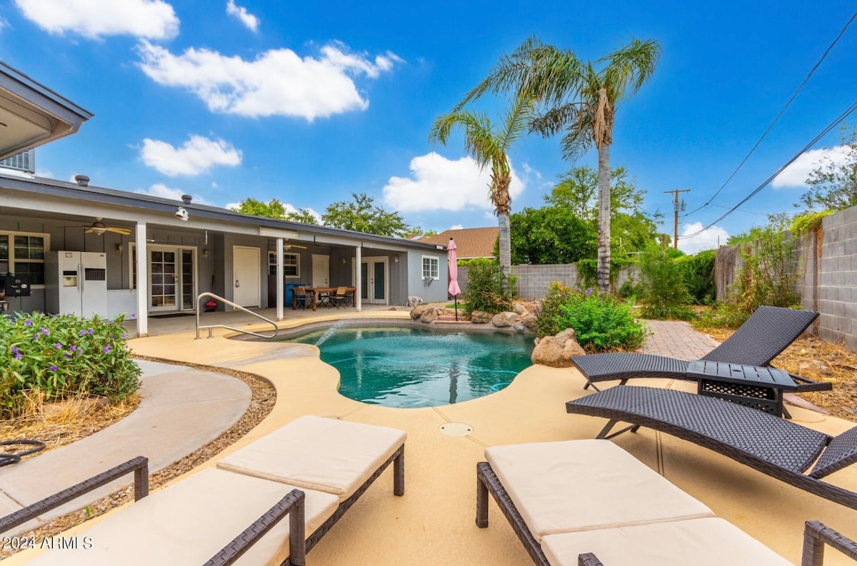 A heated and cooled play pool is surrounded by a spacious patio area with lounge chairs. Lush greenery and palm trees provide shade, while a smooth path leads to other sections of the backyard. The exterior of the home is visible in the background.