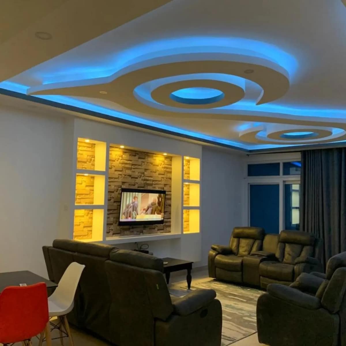 A modern living area features a sleek sectional sofa arranged for comfort, with a low table positioned in front. An entertainment unit holds a television, surrounded by decorative lighting panels. Soft, ambient light illuminates the ceiling's unique curved design, enhancing the room's contemporary feel.