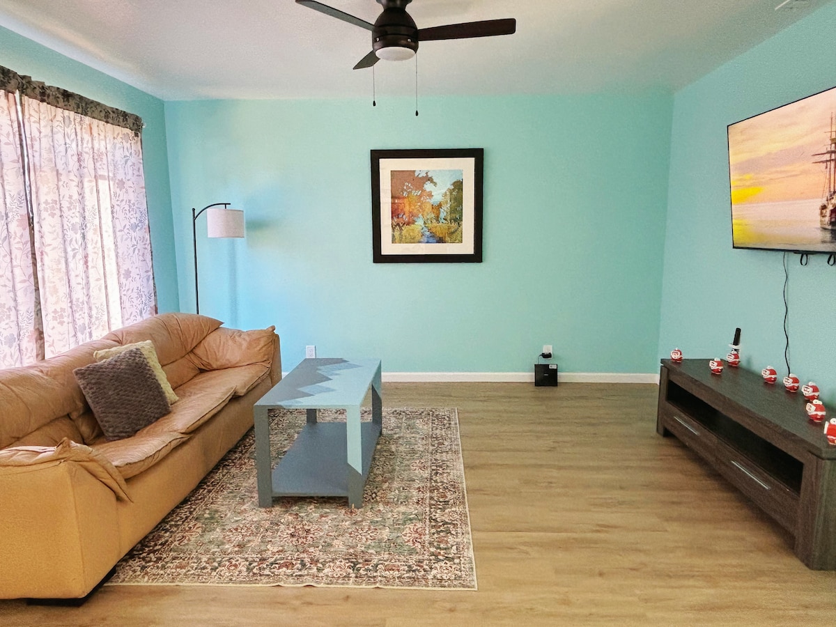 A light-filled living area features a beige couch paired with a textured throw and decorative pillows. A rectangular blue coffee table is centered on a patterned rug. The walls are painted in a calming turquoise, and large windows are dressed with sheer curtains, allowing soft light to filter in.