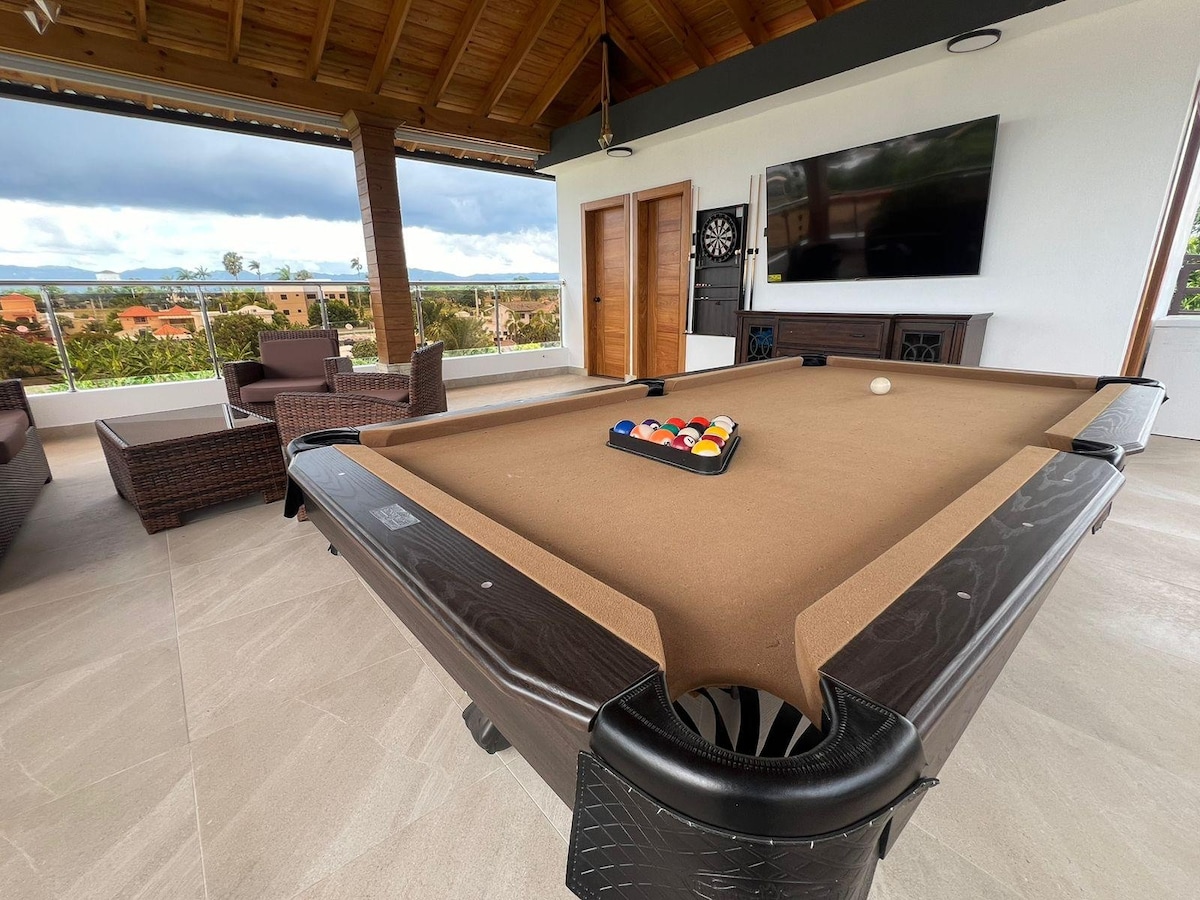 A billiard table is positioned centrally in a spacious lounge area, with a set of colorful balls arranged in a triangle. Soft seating is visible nearby, and large windows allow natural light to illuminate the space, providing views of the surrounding landscape.