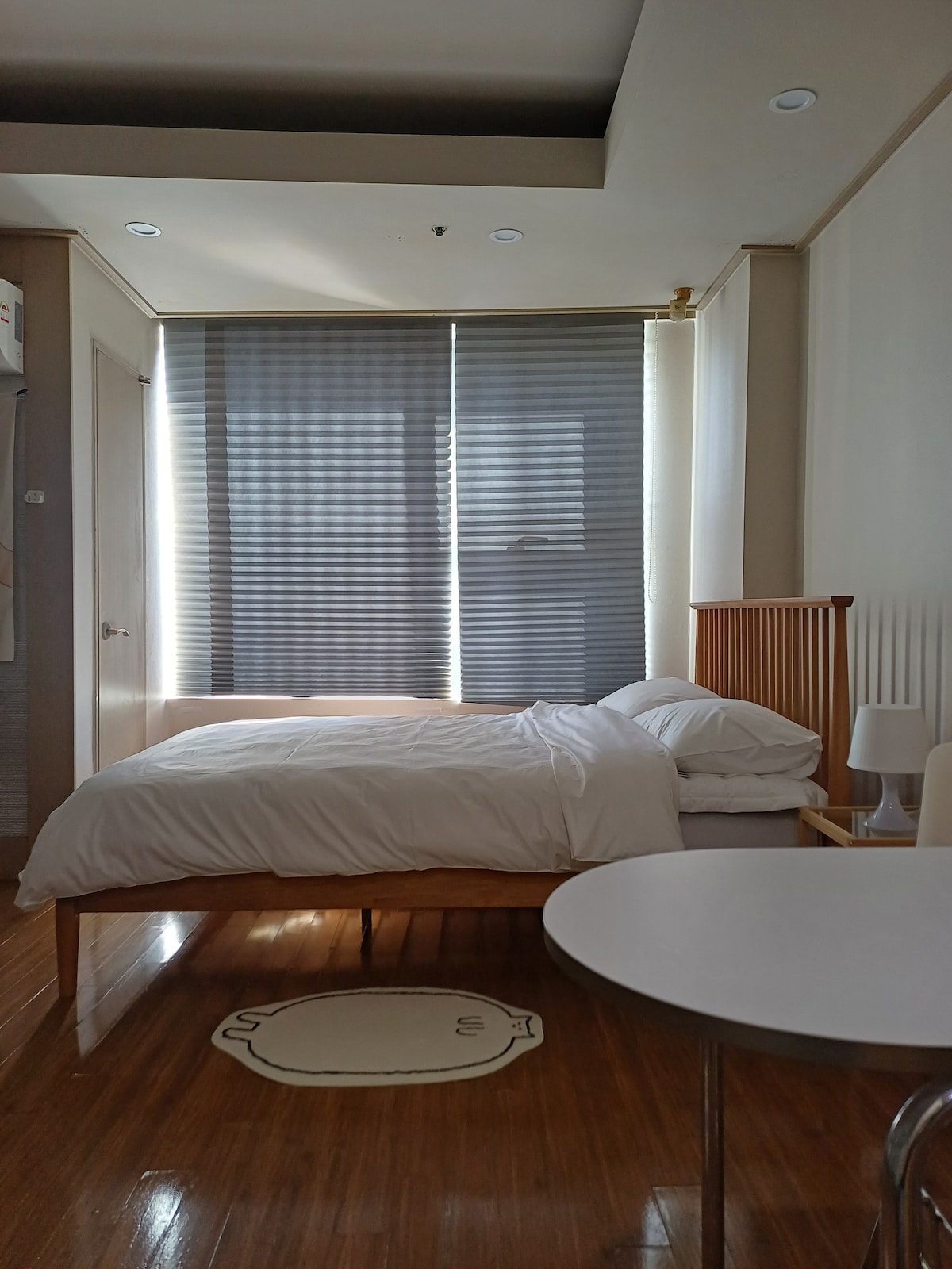A compact room features a spacious bed with white linens, positioned against a wall beside large, horizontally striped curtains that filter natural light. A small table is visible in the foreground, complementing the wooden flooring and minimalist décor.