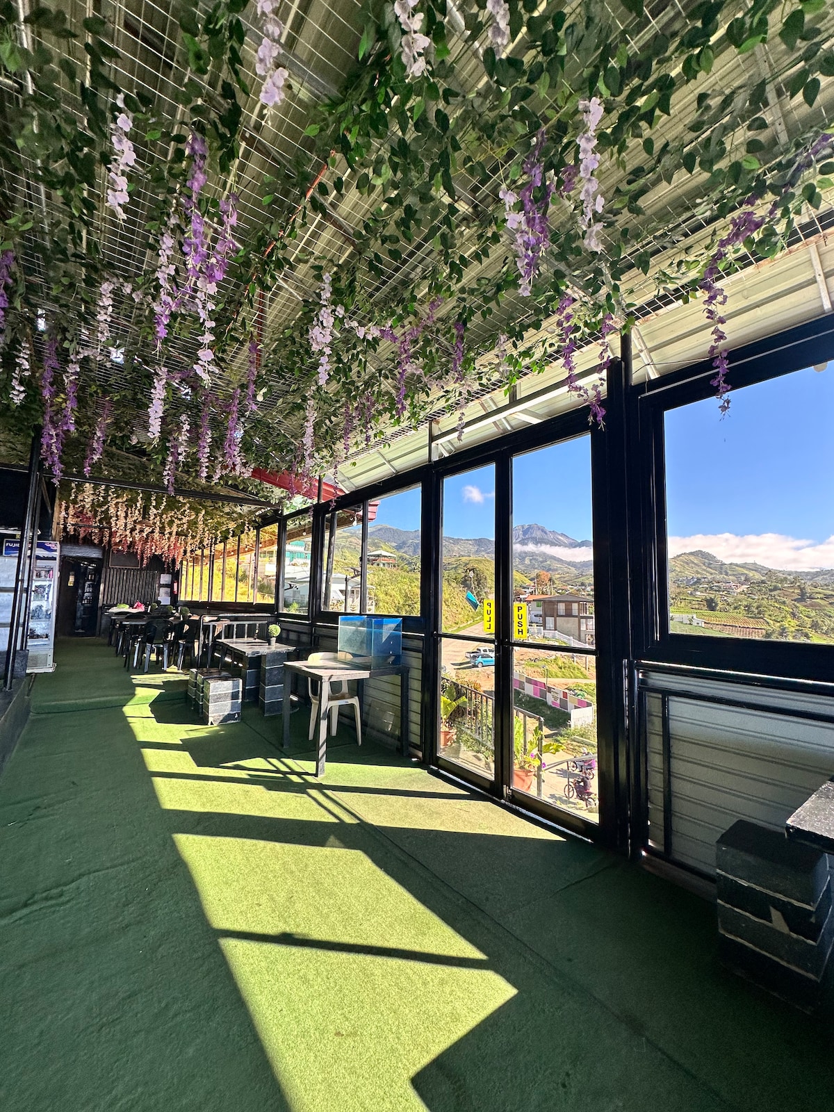 A spacious indoor area features large glass windows displaying scenic mountain views. The ceiling is adorned with hanging greenery and purple flowers. Several black tables and chairs are arranged across the carpeted floor, allowing for a comfortable and relaxed atmosphere.