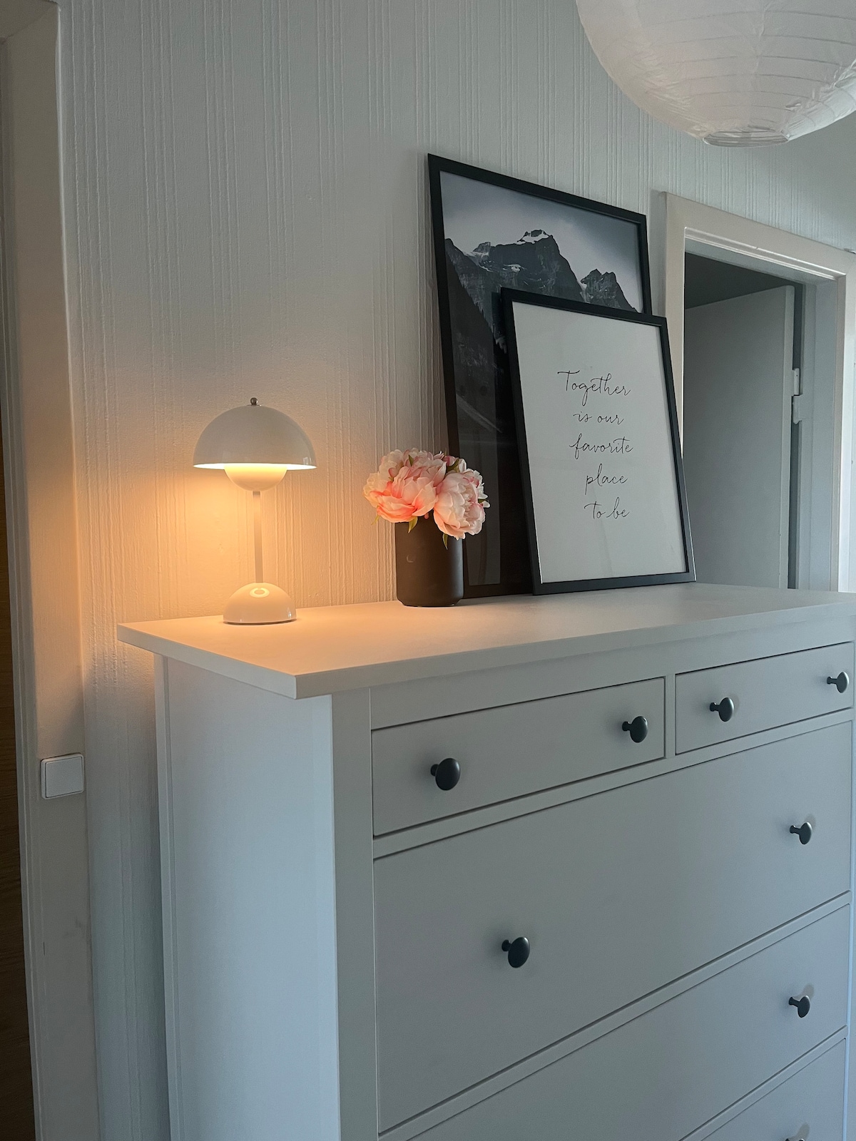 A white dresser is positioned against a light-colored wall, featuring six drawers and decorative black handles. A small lamp emits warm light, complemented by a black vase containing pink flowers. Two framed pictures, one with a mountain scene, add visual interest above the dresser.