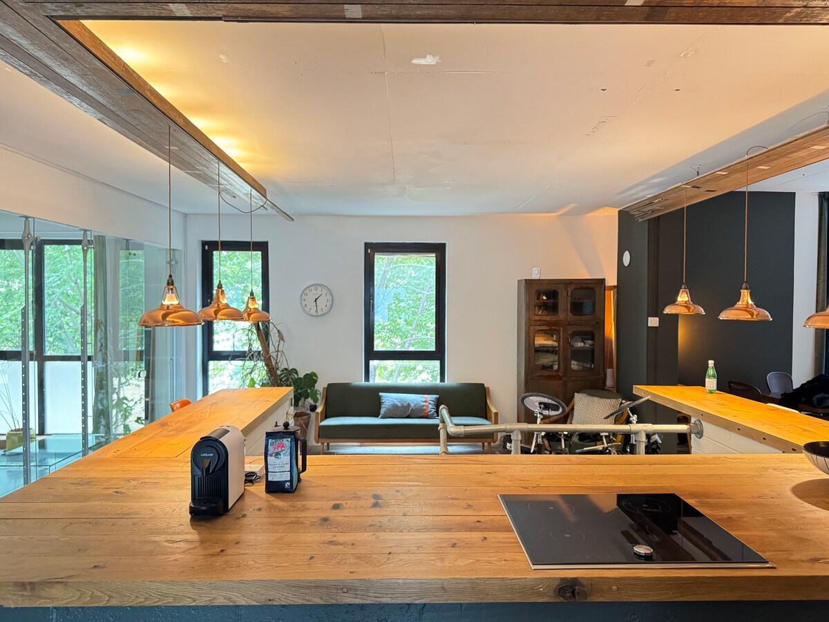 A large open-plan kitchen and dining area is visible, featuring wooden countertops, pendant lighting, and modern appliances. A green sofa is placed under a window, allowing natural light to brighten the space. Decorative plants contribute to the inviting atmosphere.