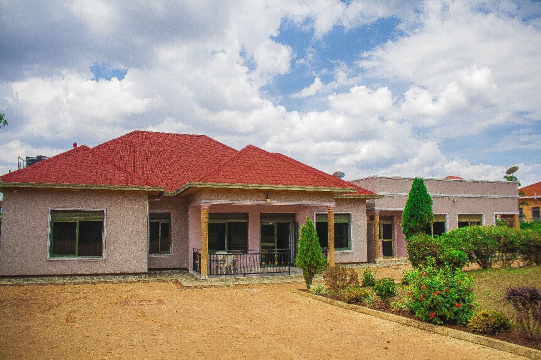 A spacious house is presented with a red-tiled roof and neutral-colored exterior walls. Lush green shrubs and trees surround the property, while a gravel driveway leads up to the entrance. Large windows provide natural light to the living areas.