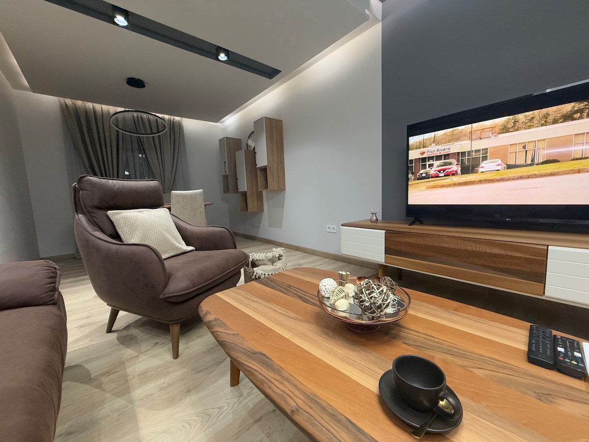 A modern living area features a plush armchair and a streamlined wooden coffee table arranged in front of a flat-screen television. Neutral tones dominate the decor, complemented by soft lighting and an accent wall. Decorative items are displayed on the table, enhancing the inviting ambiance.