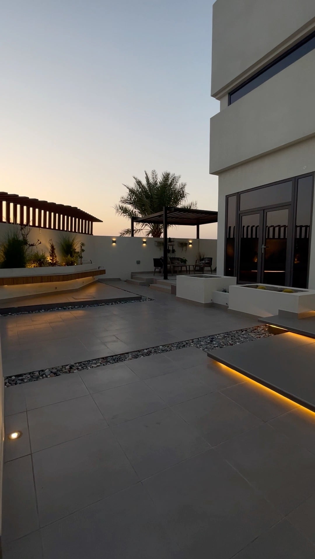 An outdoor area features a modern design with sleek tiles and integrated lighting along pathways. A tranquil swimming pool is set against the backdrop of an evening sky. Lush greenery adds to the serene ambiance, complemented by a covered seating area under a pergola.