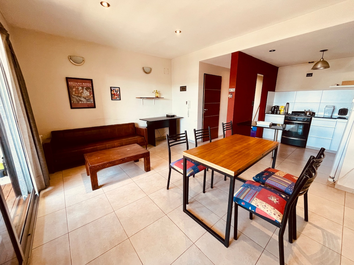 An open-plan living area features a wooden dining table surrounded by chairs, with a sofa positioned against the wall. A kitchenette is visible with modern appliances, while large windows allow ample natural light to fill the space.