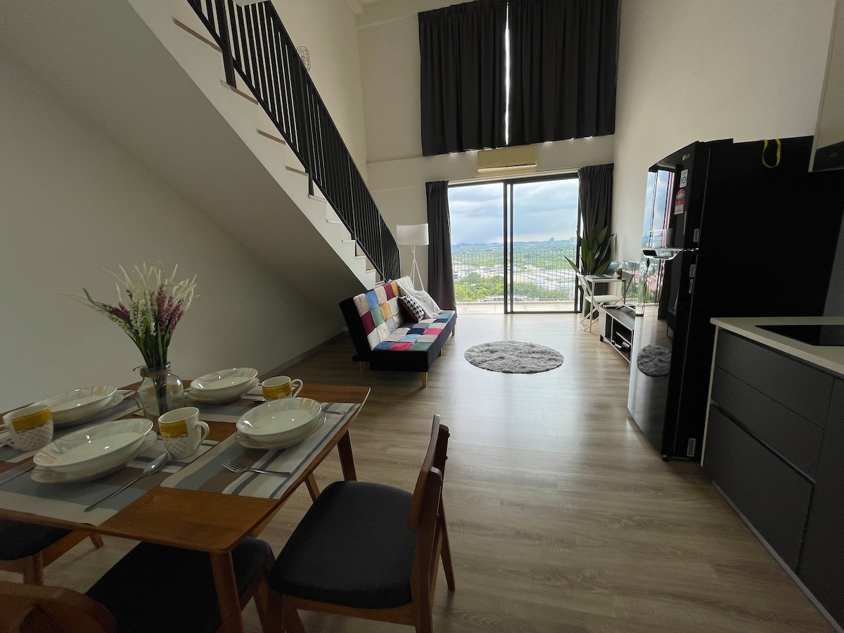 An open living area features a comfortable sofa adorned with colorful cushions, paired with a dining table set for four. Large windows allow natural light to fill the space, while a kitchenette and a rug add to the cozy atmosphere. A staircase leads to the upper level.