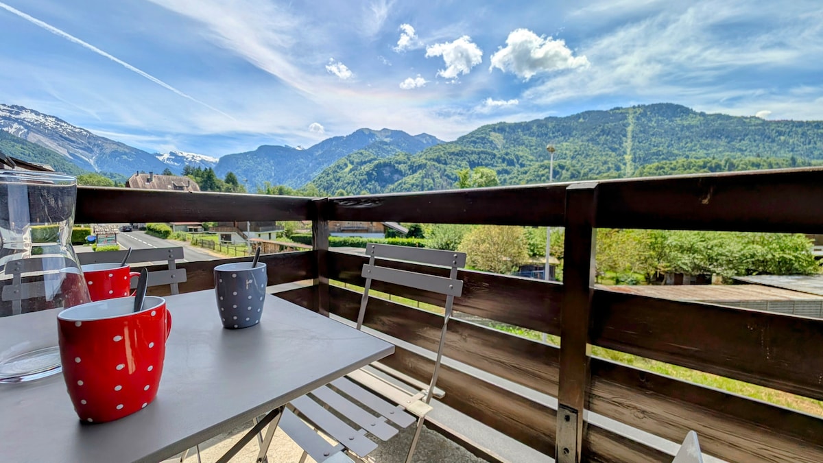 A balcony scene showcases a table with two coffee mugs, one in red with white polka dots and the other in grey. The mountains rise in the background under a clear blue sky, providing a serene outdoor setting for relaxation.