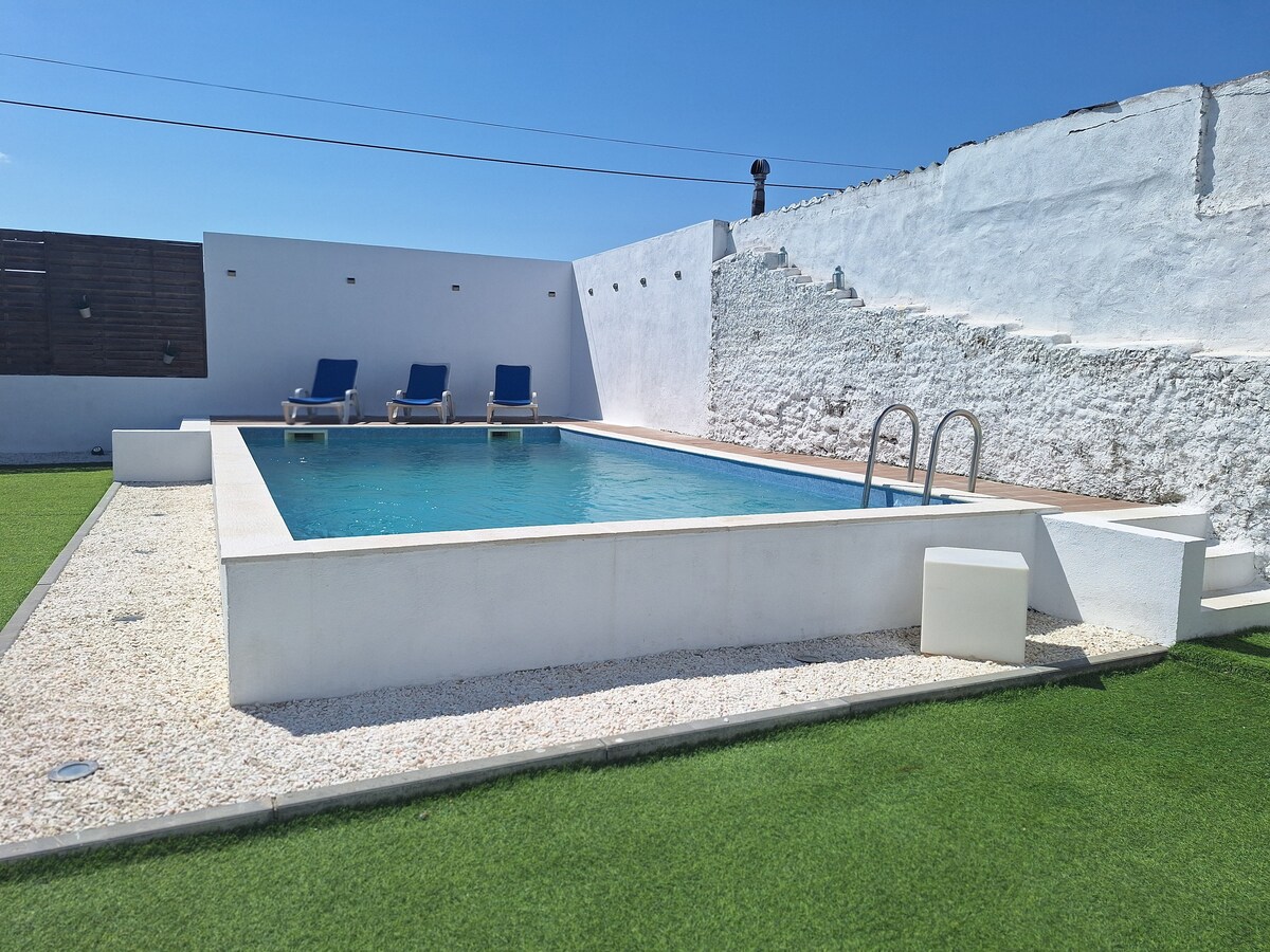 A rectangular pool is surrounded by artificial grass, providing a neat and inviting area. Several lounge chairs are positioned at one end of the pool deck, set against a backdrop of a white wall reflecting the sunlight. Clear blue skies enhance the vibrant setting.