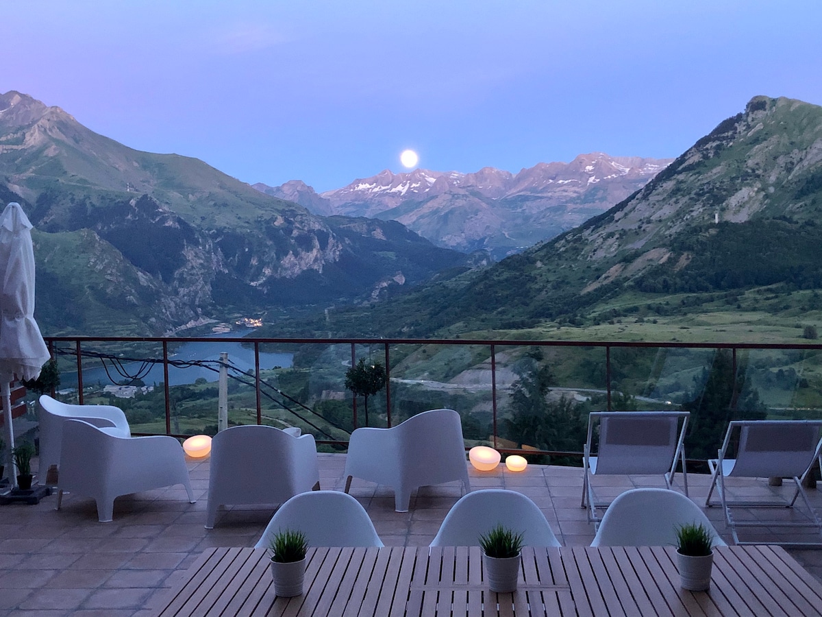 A spacious terrace offers stunning views of rolling mountains and a serene lake. Several white lounge chairs are arranged around a wooden table, while soft lighting creates a peaceful ambiance. A full moon rises over the landscape, enhancing the tranquility of the setting.
