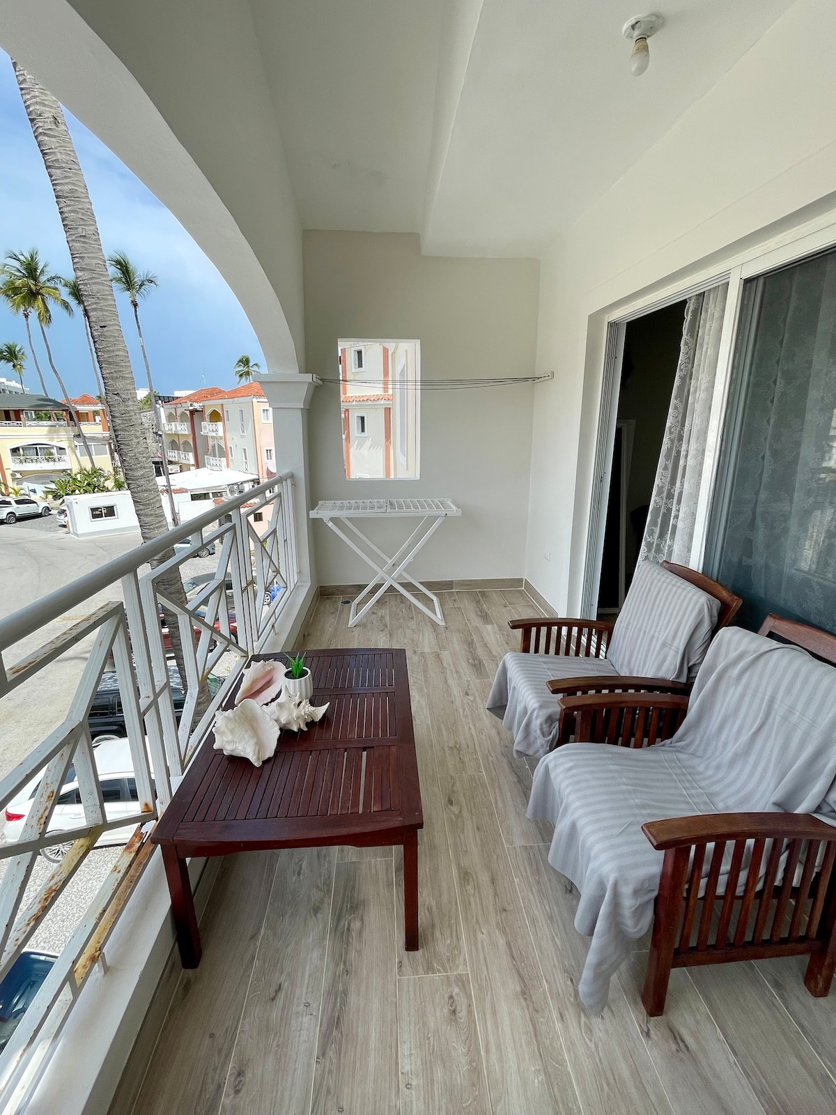 A balcony area is furnished with two wooden armchairs and a small table. An airer is positioned nearby, and the space is surrounded by palm trees, offering a view of the bustling area below. Light-colored walls complement the outdoor setting.