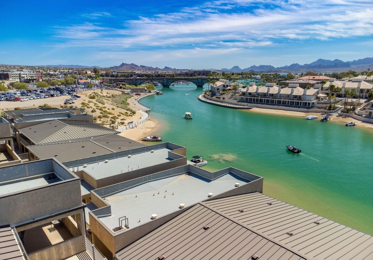 Aerial view of the sparkling green waterway, surrounded by sandy shores and palm trees. Nearby buildings with flat roofs are visible, along with boats gliding through the water under a clear blue sky, showcasing the vibrant atmosphere of the area.