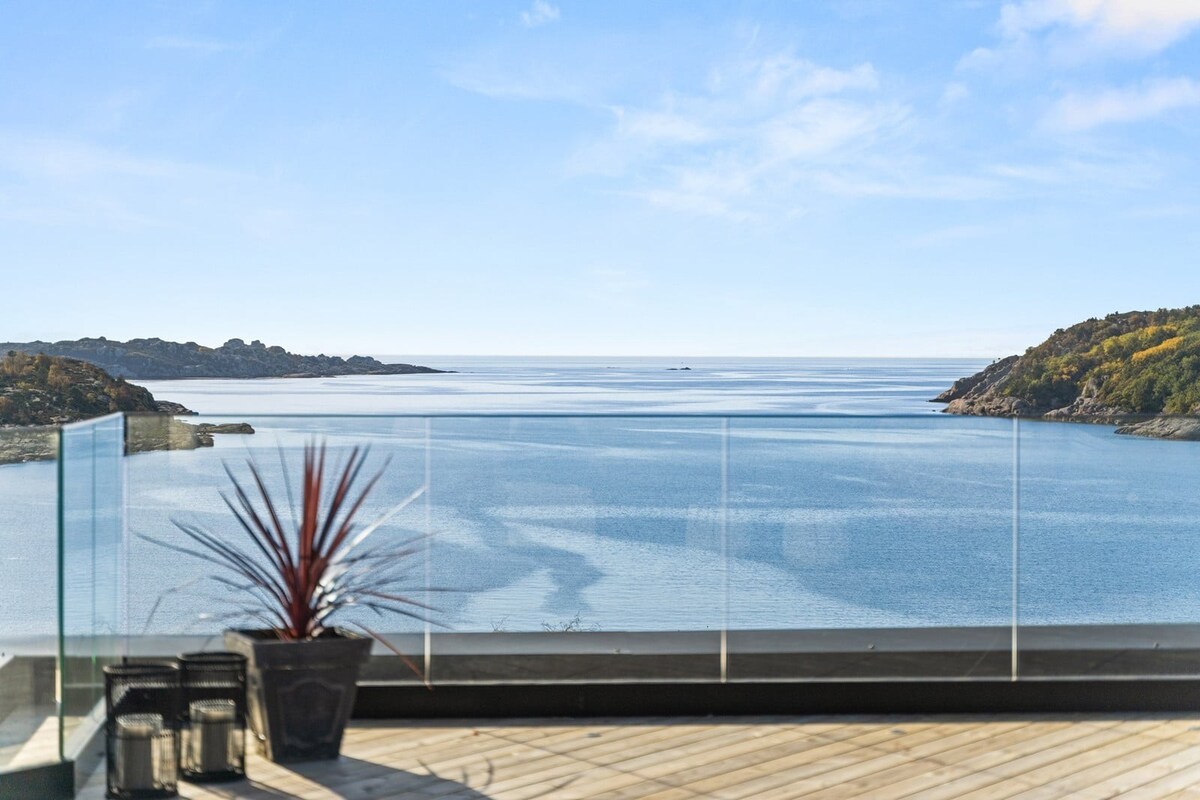 A panoramic view of the water is framed by a clear glass railing. A potted plant adds a touch of greenery to the foreground. The horizon features gentle hills, while the serene waters reflect the sky's colors, creating a tranquil setting.
