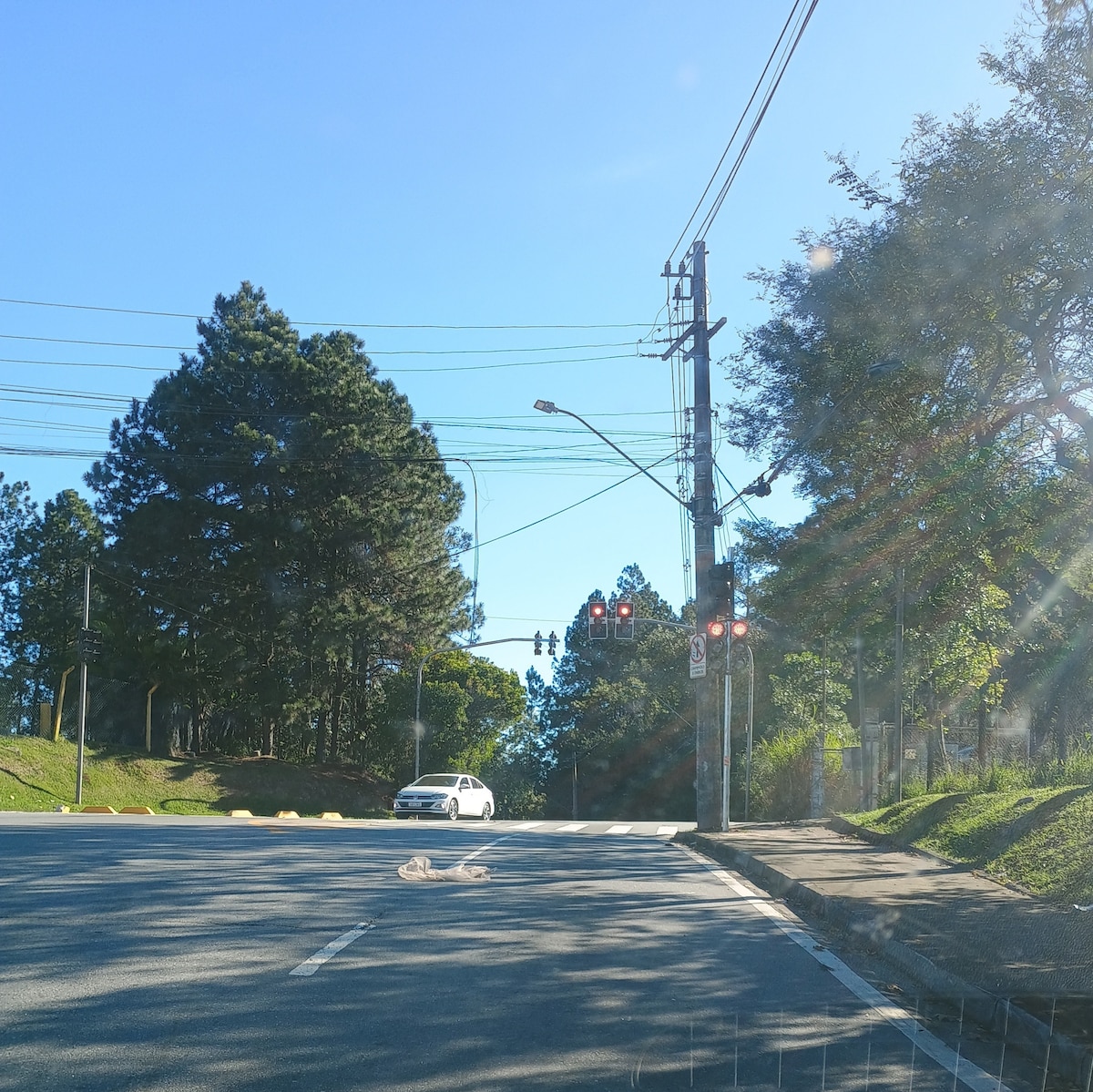 A clear view of a quiet intersection is shown, with traffic lights displaying red signals. Lush green trees line the road, basking in sunlight. A single vehicle is parked at the intersection, indicating the presence of a peaceful and orderly environment.