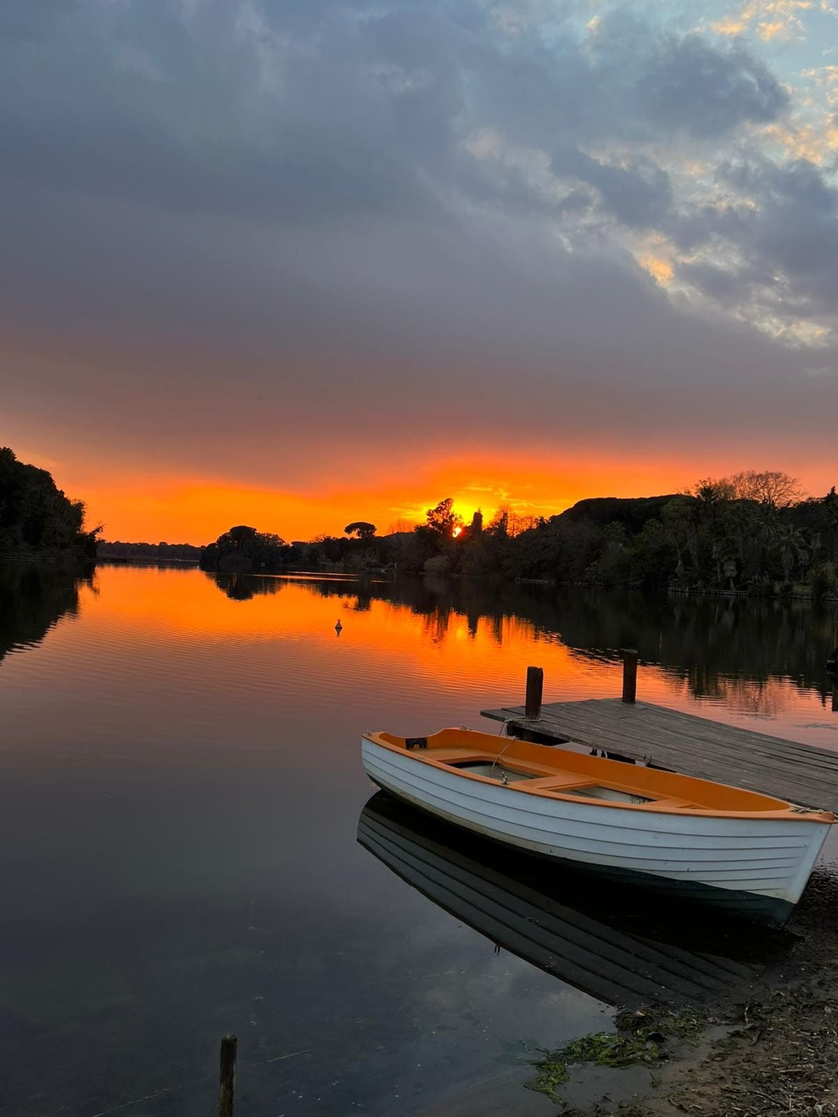 A serene body of water reflects vibrant orange and yellow hues from a sunset, framed by trees along the bank. A small white rowing boat with an orange interior rests on a wooden dock, adding to the tranquil atmosphere. Light clouds are visible in the sky.