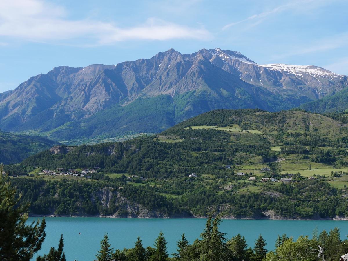 The image showcases a panoramic view of mountains with snow-capped peaks and lush green slopes, set against a bright blue sky. A serene lake is visible at the foothill, surrounded by forested areas, creating a harmonious natural landscape.