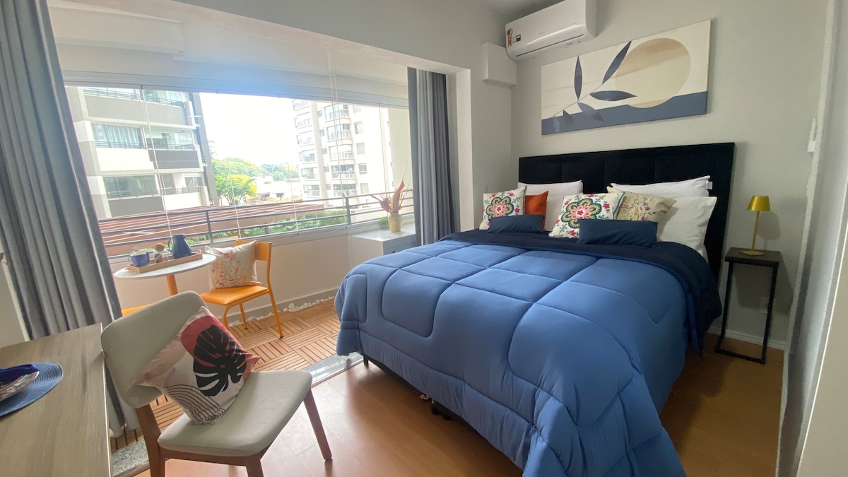 A cozy bedroom is furnished with a spacious bed covered by a blue comforter. Decorative pillows add a pop of color, while natural light filters through large windows with sheer curtains. A small dining table with two chairs is positioned nearby, offering a pleasant view.
