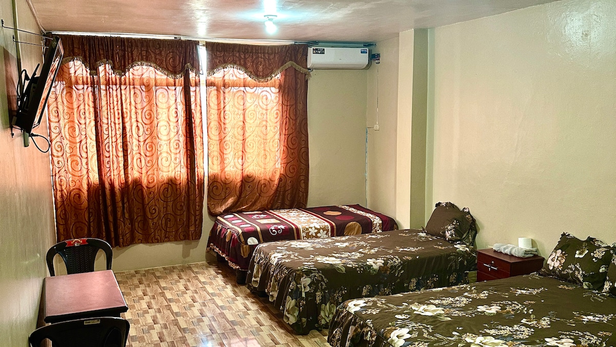 The room features three beds with floral-patterned linens, providing ample sleeping space. A large window is covered by sheer brown curtains, allowing natural light to filter in. A small wooden table is placed against the wall, complemented by a TV mounted above.