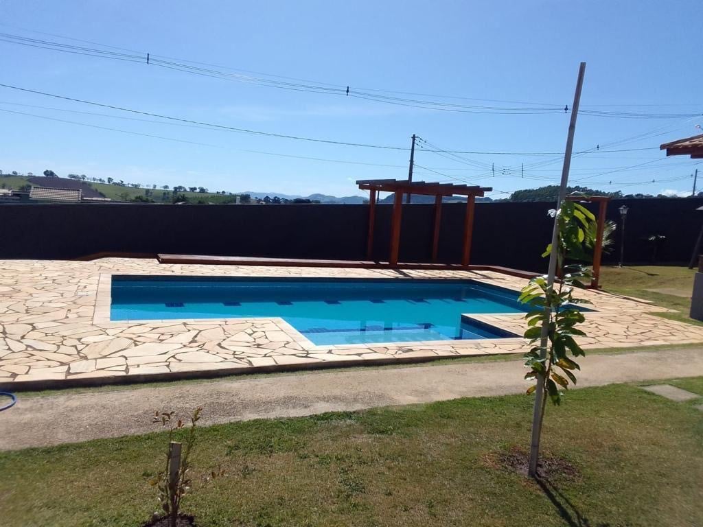 An outdoor area includes a clear swimming pool framed by a stone patio. A pergola is situated beside the pool, and lush green grass surrounds the space. Scenic hills can be seen in the background under a bright blue sky.