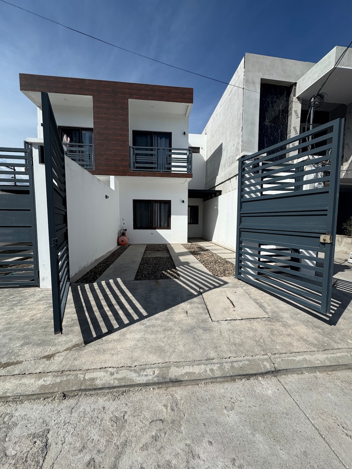 The entrance features modern metal gates, which are opened to reveal a pathway leading to a two-story building. The area is surrounded by a mix of textures, including smooth concrete and gravel. Natural sunlight creates shadows on the ground, enhancing the contemporary design.