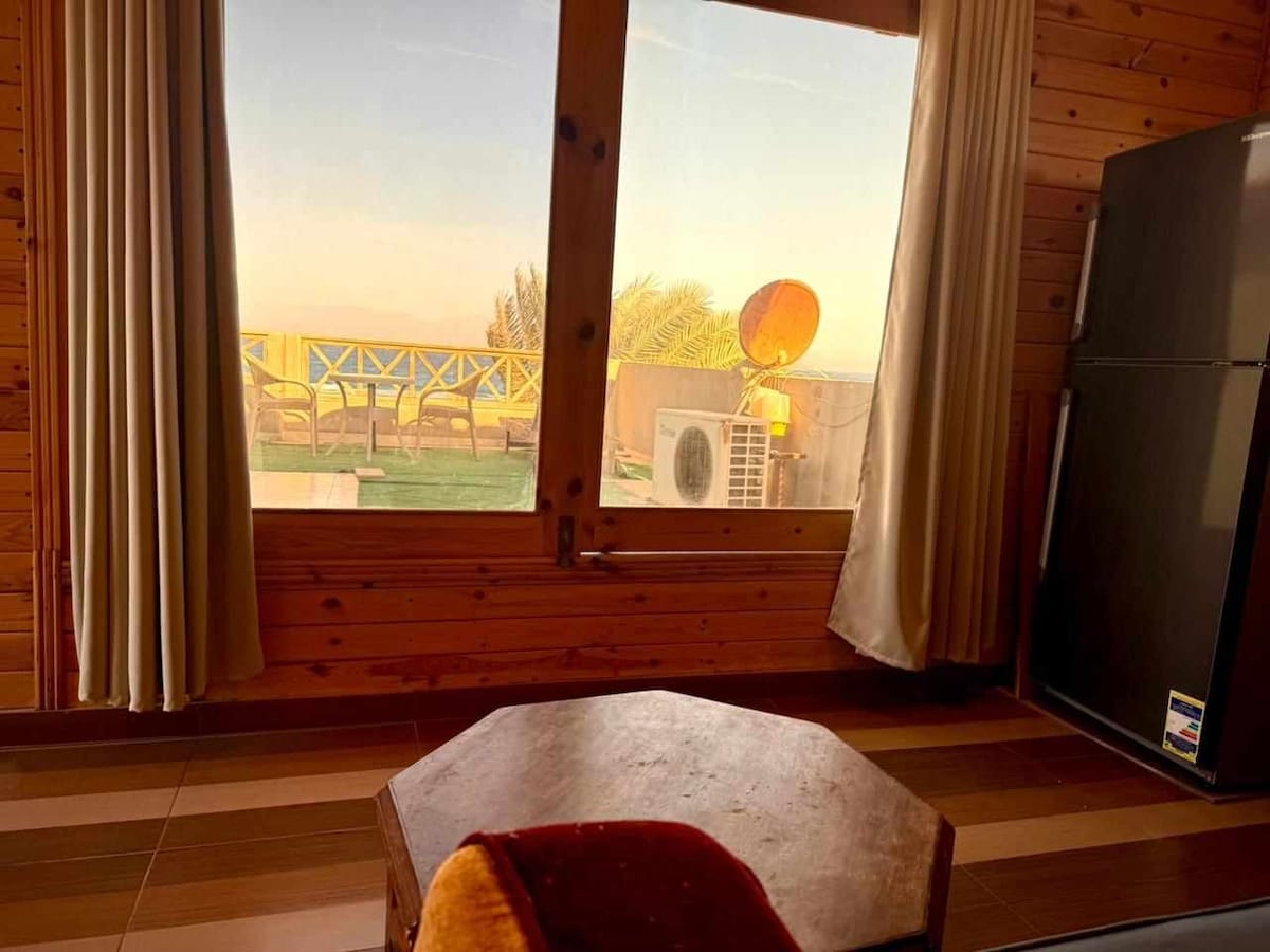 A large window frames a view of outdoor seating and greenery. Natural light illuminates the room, highlighting a wooden table and a refrigerator positioned against the wall. Light-colored curtains frame the window, complementing the warm wooden paneling.