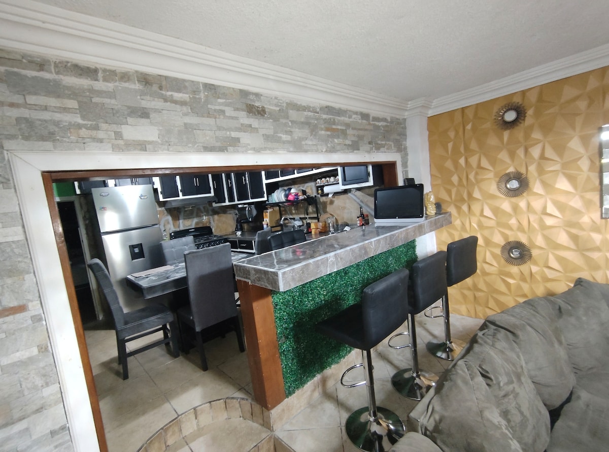 An open-concept kitchen and dining area showcase modern amenities. A sleek bar counter with high stools is positioned beside a dining table. Textured wall finishes in warm hues add depth to the space, while appliances and decorative elements reflect a contemporary style.