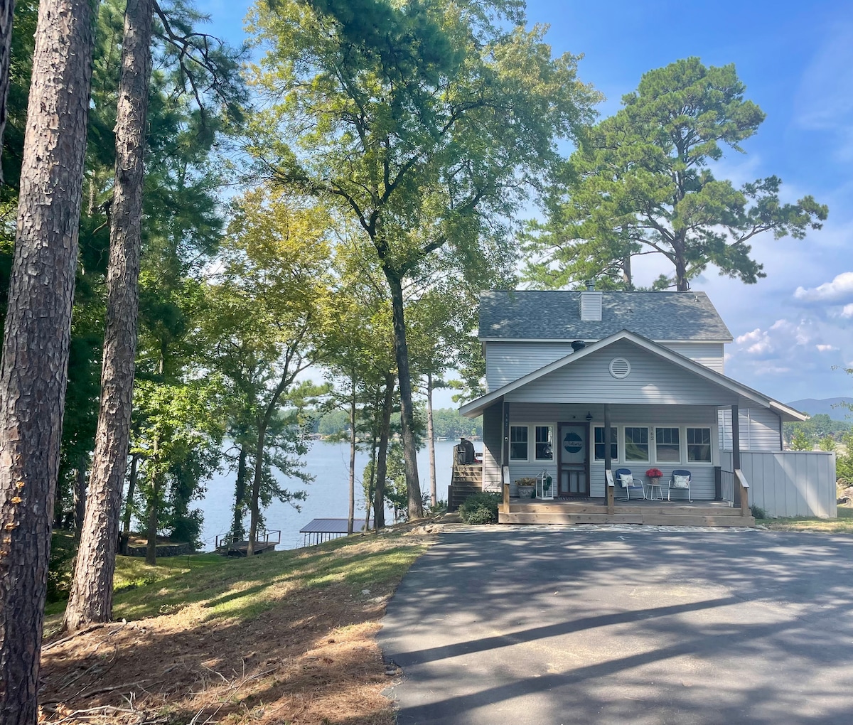 A charming, two-story building is nestled among towering trees, with a welcoming porch overlooking the lake. The exterior features a light grey façade, and a pathway leads to the water's edge, inviting exploration and relaxation in a natural setting.