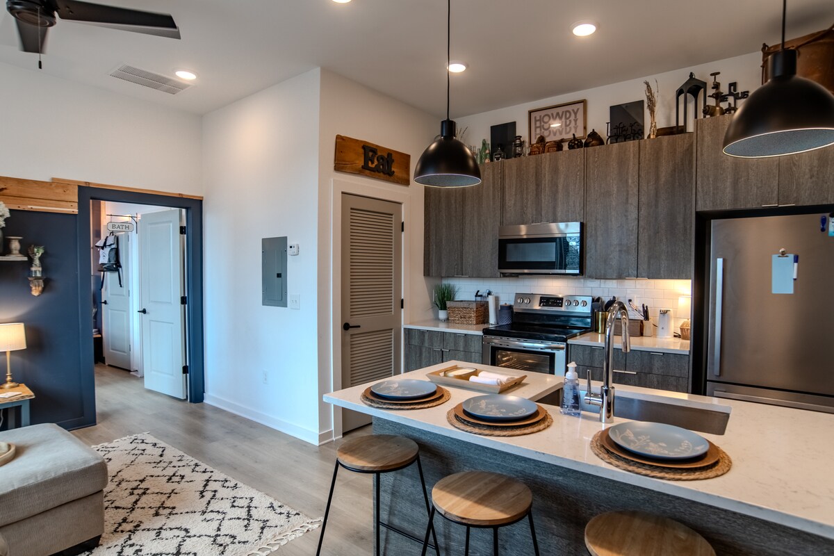 An open kitchen area features sleek cabinetry and modern appliances. A breakfast bar with three wooden stools provides seating. Warm lighting illuminates the space, while a cozy living area is visible in the background, showcasing a neutral color palette and a textured rug.