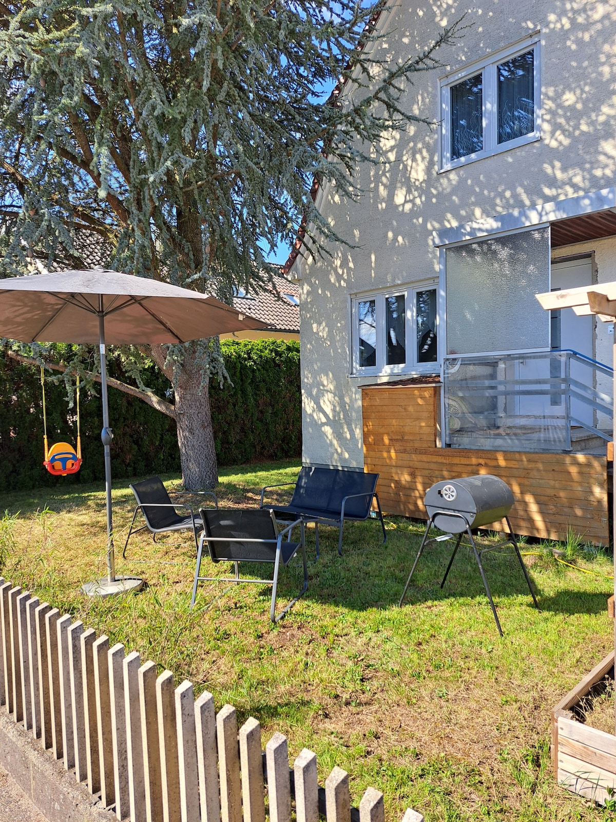 A shaded outdoor area features a patio set comprising two chairs and a small table, complemented by a grill and a swing. A large umbrella provides protection from the sun, while a tree offers additional shade. The grass is well-kept, enhancing the space's relaxed ambiance.