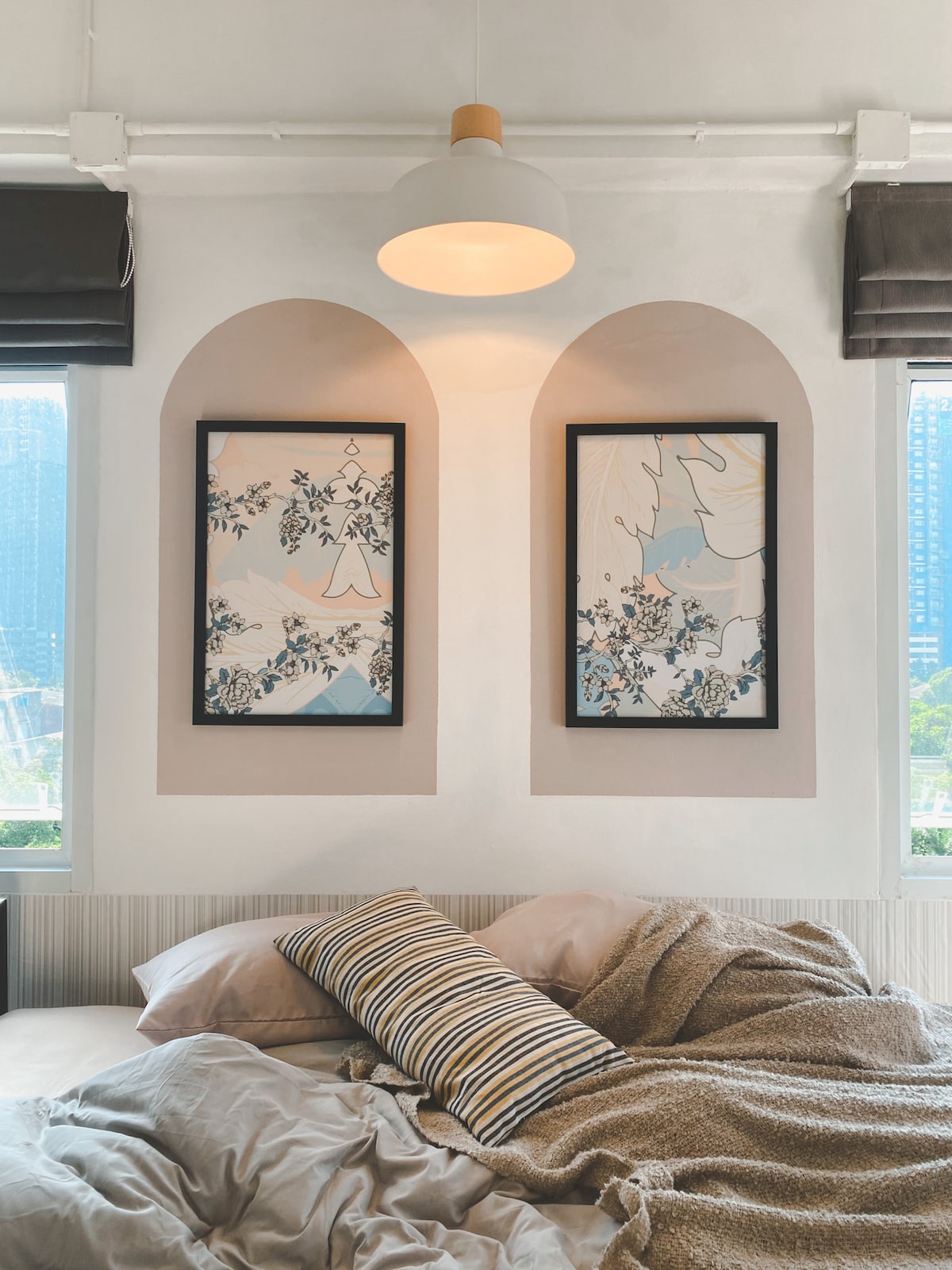 A cozy sleeping area features soft bedding with neutral tones. Two framed artworks are positioned over the bed, showcasing detailed botanical designs. The room is illuminated by a ceiling light, and window coverings allow natural light to filter in, creating a serene environment.