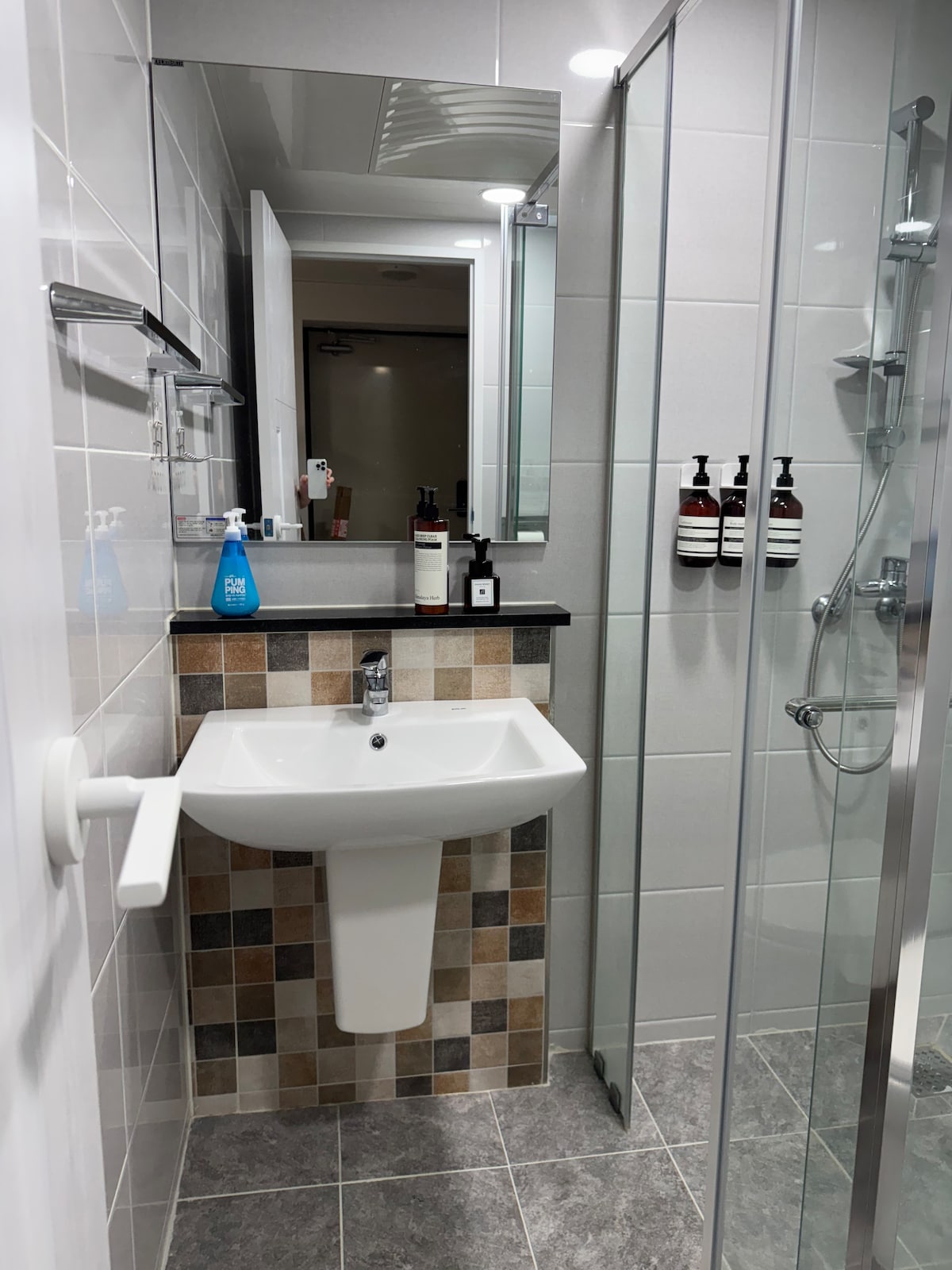 A modern bathroom is shown, featuring a glass shower and a stylish sink mounted on a tiled wall with varying colors. Toiletries are neatly arranged on a shelf, and bright lighting accents the clean, contemporary design.