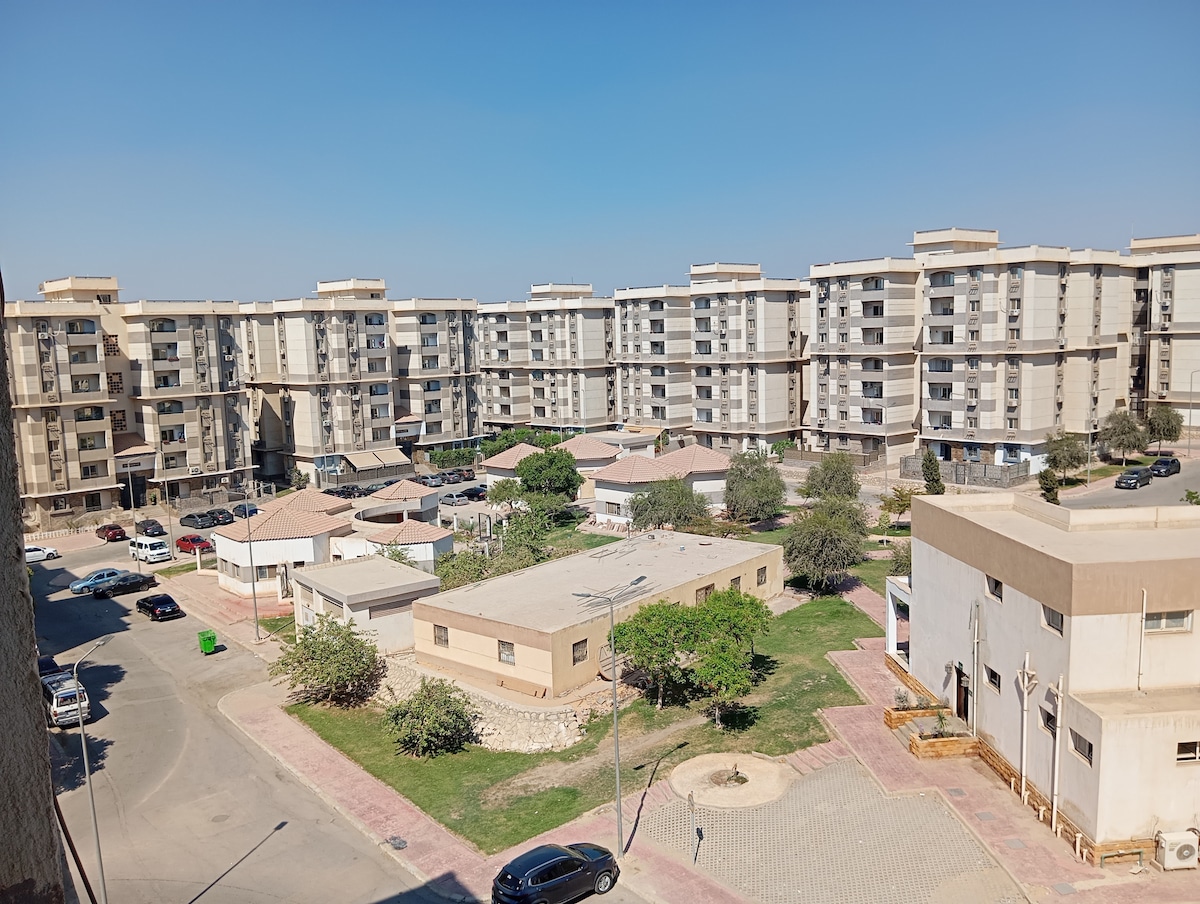 A view of the residential complex features multiple buildings arranged around a green space. Pathways are visible, leading to shaded areas with seating. The surrounding landscape includes trees and well-maintained lawns, contributing to a serene environment. Cars are parked in designated areas, indicating accessibility.