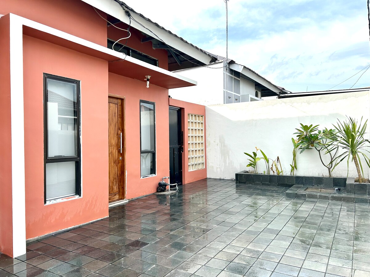 A modern exterior of the vacation home features a vibrant coral-colored wall and large windows, providing natural light. A spacious tiled patio is seen, bordered by low-maintenance greenery, enhancing the outdoor living space. A simple privacy wall separates the outdoor area from the surroundings.