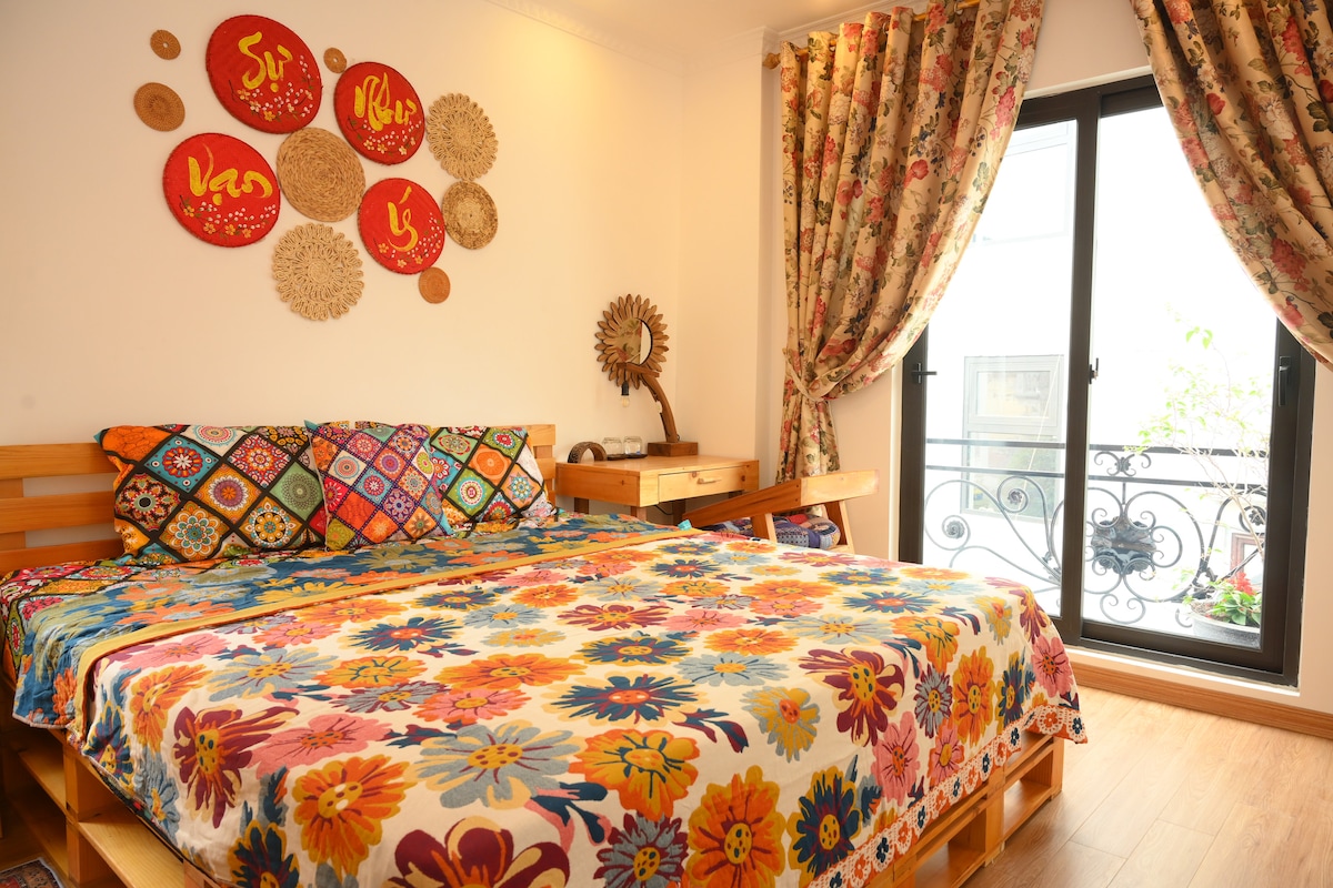 A vibrant bedroom features a spacious bed adorned with a colorful floral blanket. Decorative wall art and textiles add warmth to the space. Natural light enters through a glass door, which leads to a small balcony, while patterned curtains frame the window.